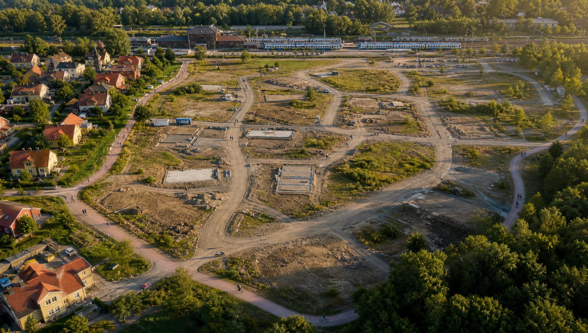 Fågelvy över ett tidigt skede av markutveckling – obebyggda tomter, grundarbeten och färdiga hus längs kanten, med tågstation i bakgrunden